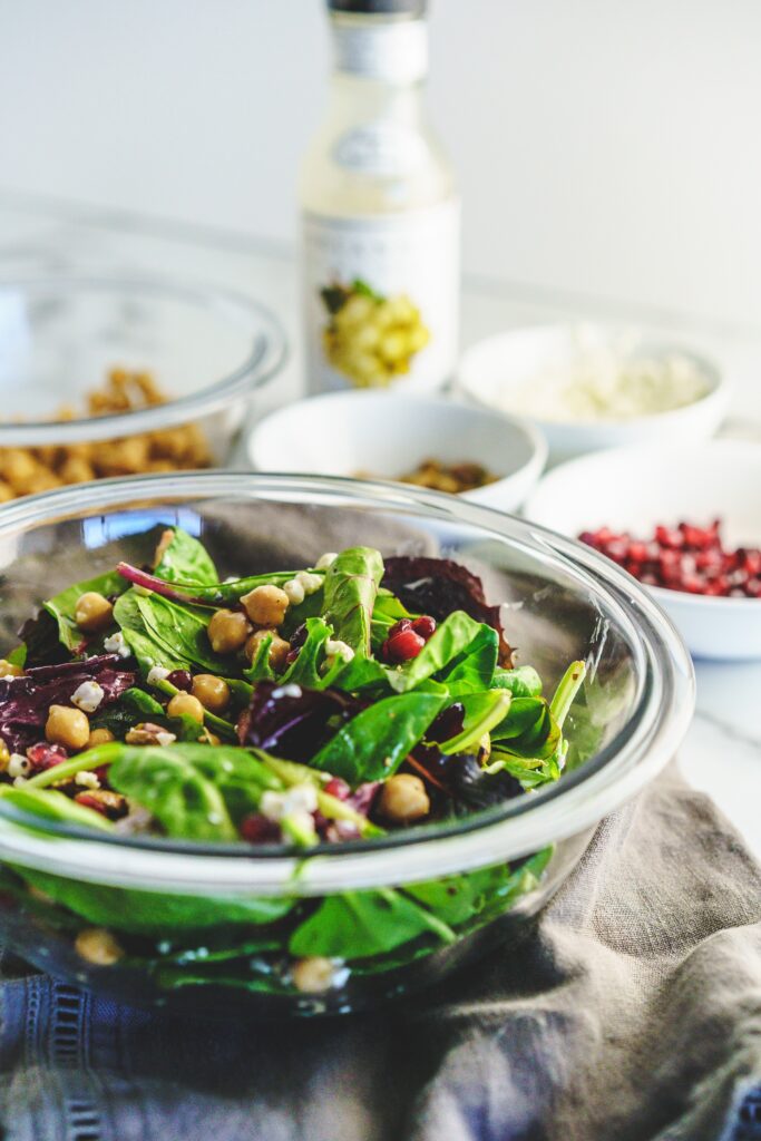 clear bowl of mixed greens topped with chickpeas, pistachios, goat cheese, and pomegranate arils