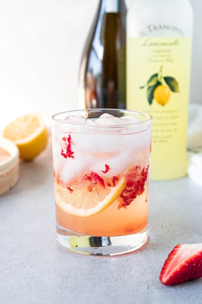 glass filled with ice, strawberry pieces, cocktail, and lemon slice with bottles in background