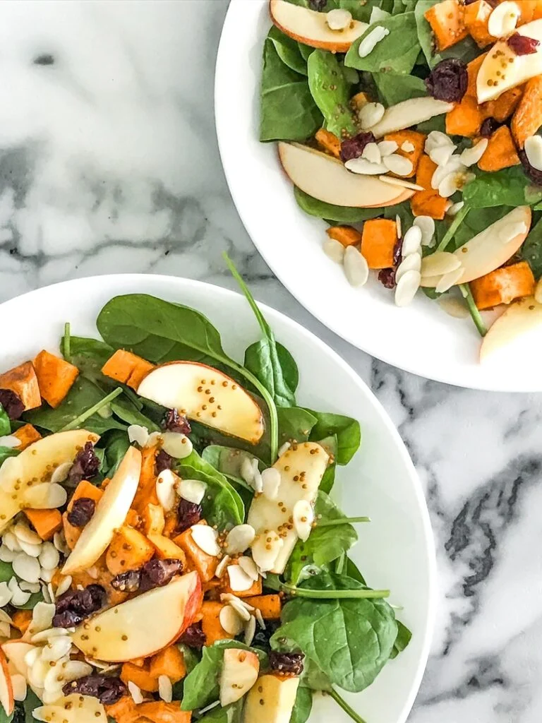 white plate with spinach salad topped with apples, cranberries, almonds, and sweet potatoes