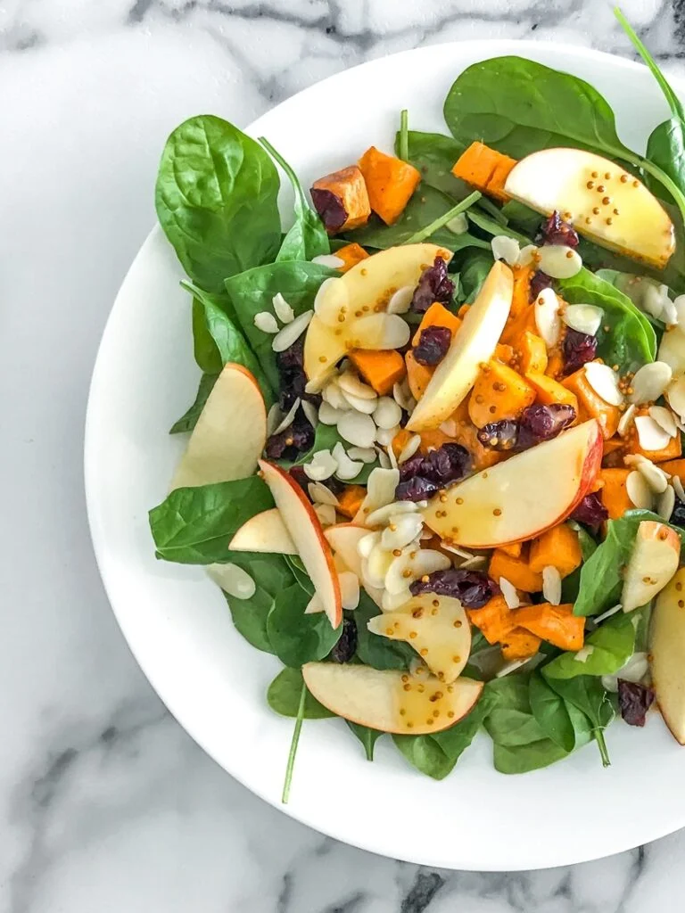white plate with spinach salad topped with apples, cranberries, almonds, and sweet potatoes