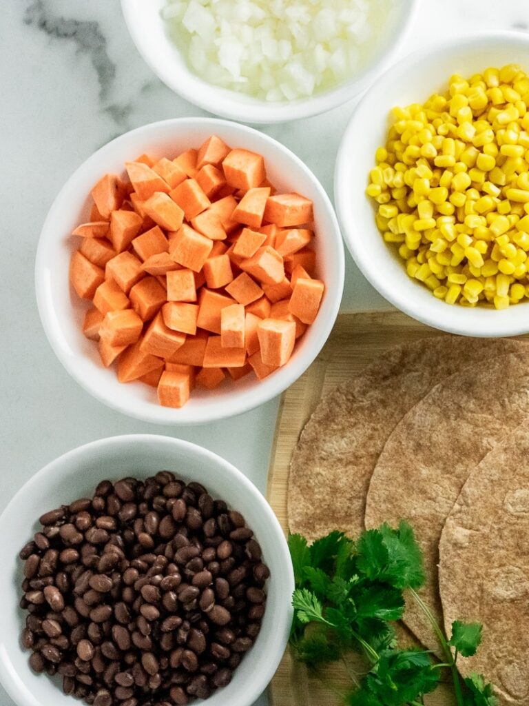 white bowls with black beans, sweet potatoes, and corn next to cutting board and tortillas