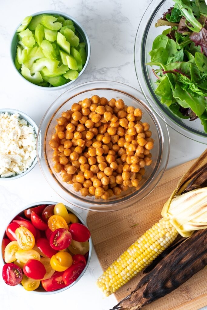 ear of corn and bowls with feta cheese, celery, tomatoes, chickpeas, and greens