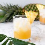 glass with pineapple juice, pineapple wedge, and ice with pineapple in background