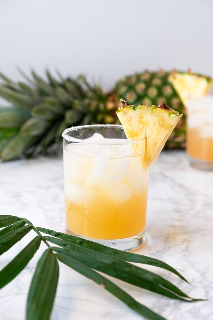 glass with pineapple juice, pineapple wedge, and ice with pineapple in background