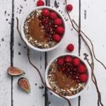 two chocolate smoothie bowls topped with chopped nuts, pomegranate arils, and raspberries