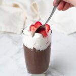 spoon digging into cup of chocolate mousse topped with whipped cream and strawberries