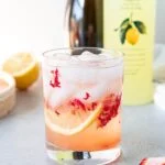 glass filled with ice, strawberry pieces, cocktail, and lemon slice with bottles in background
