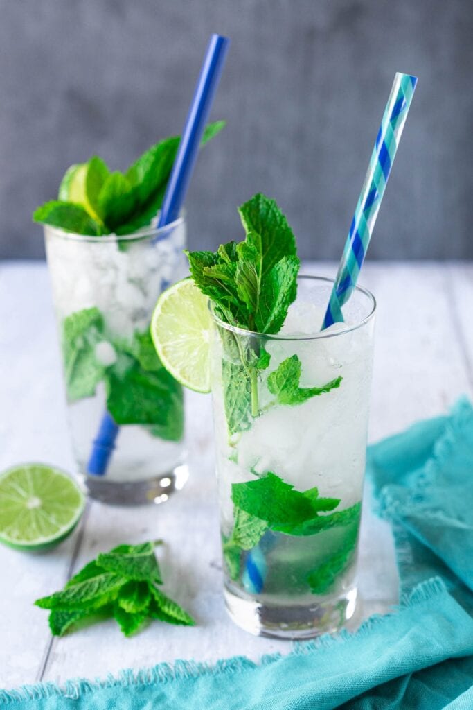 two highball glasses filled with crushed ice, water, and mint, topped with colorful straws, lime, and mint