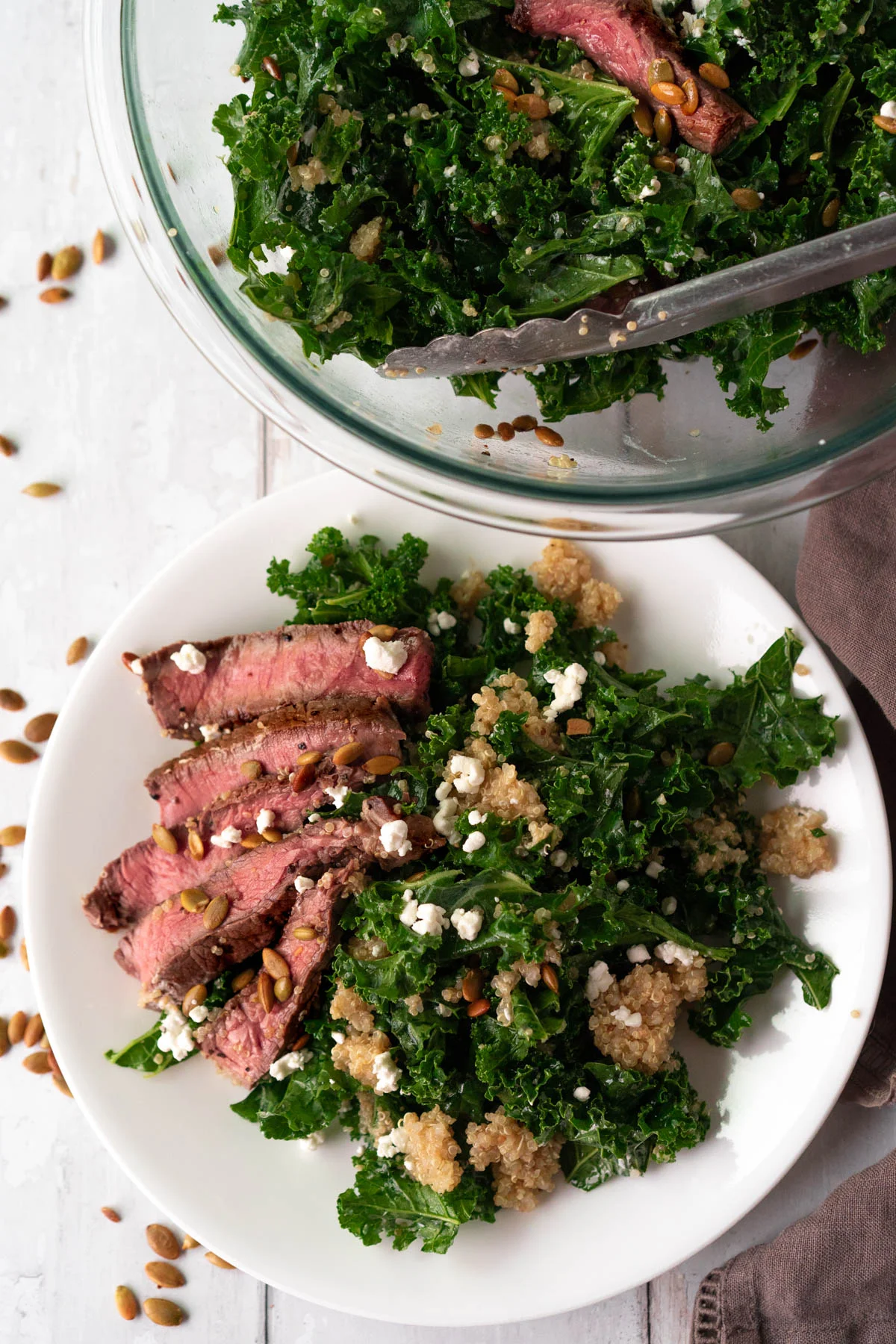 white plate with a kale salad and slices steak and large glass bowl with mixed salad and tongs in the background