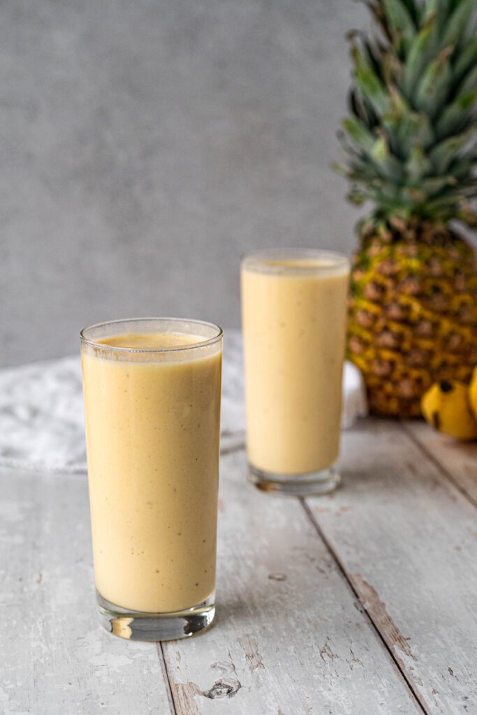 two glasses filled with a pale yellow smoothie and a pineapple in the background