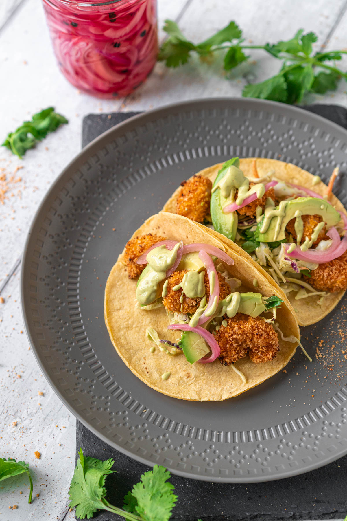 grey plate with two tacos filled with breaded seafood, avocado, and pickled onions