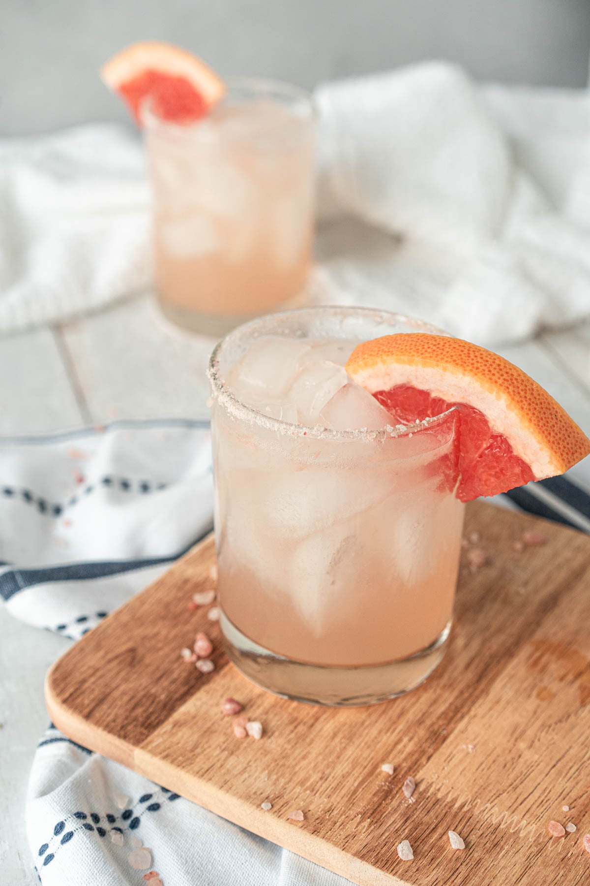 In the foreground, rocks glass sitting on a wood cutting board, glass is filled with light pink drink and garnished with pink salt and a grapefruit wedge. Blurred in the background, a matching glass is sitting against a white and grey backdrop