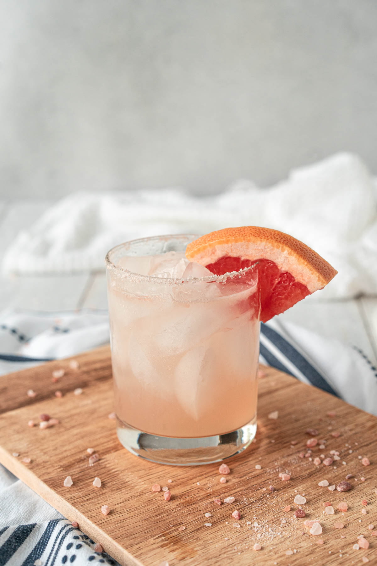 Rocks glass filled with a peach-colored drink against a grey and white background. Rim of the glass is garnished with pink salt and a grapefruit wedge. Glass is sitting on a wooden cutting board with pink salt crystals scattered across it.