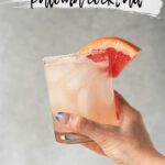 hand holding a rocks glass filled with a pale pink drink garnished with a grapefruit wedge and rimmed with salt