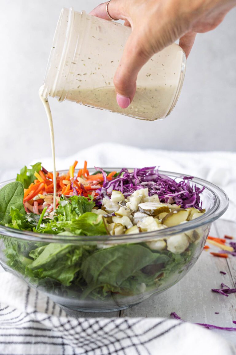 hand holding jar of ranch dressing and pouring it over a large bowl of salad with lettuce, shredded carrots, shredded cabbage, and chopped pickles