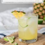 In the foreground, a rocks glass filled with ice and a light yellow drink, garnished with lime and pineapple wedges sits on a cutting board. Blurred in the background is a large pineapple and sliced limes