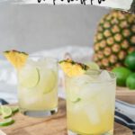 In the foreground, a rocks glass filled with ice and a light yellow drink, garnished with lime and pineapple wedges sits on a cutting board. Blurred in the background is a large pineapple and sliced limes