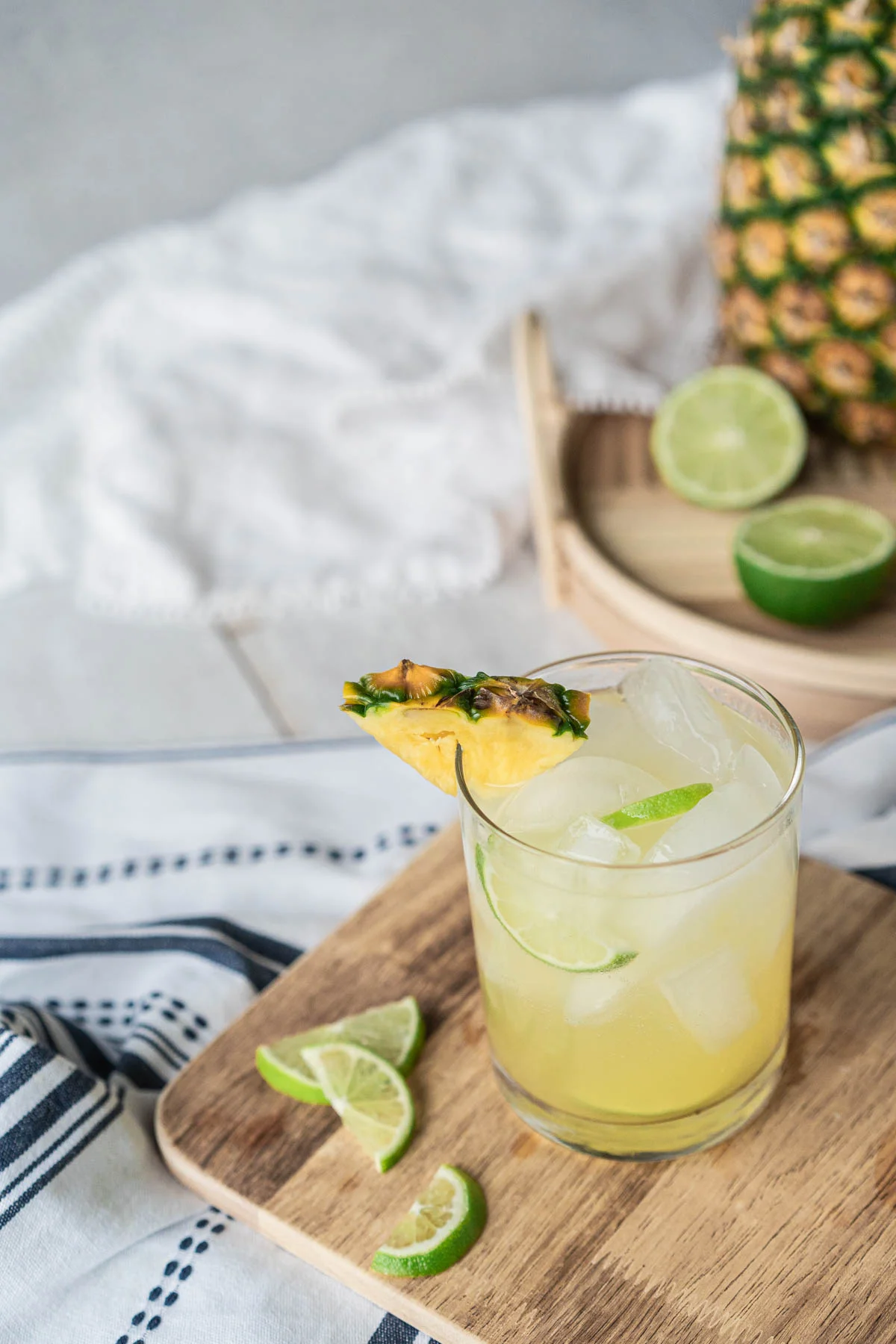 In the foreground, a rocks glass filled with ice and a light yellow drink, garnished with lime and pineapple wedges sits on a cutting board. Blurred in the background is a large pineapple and sliced limes