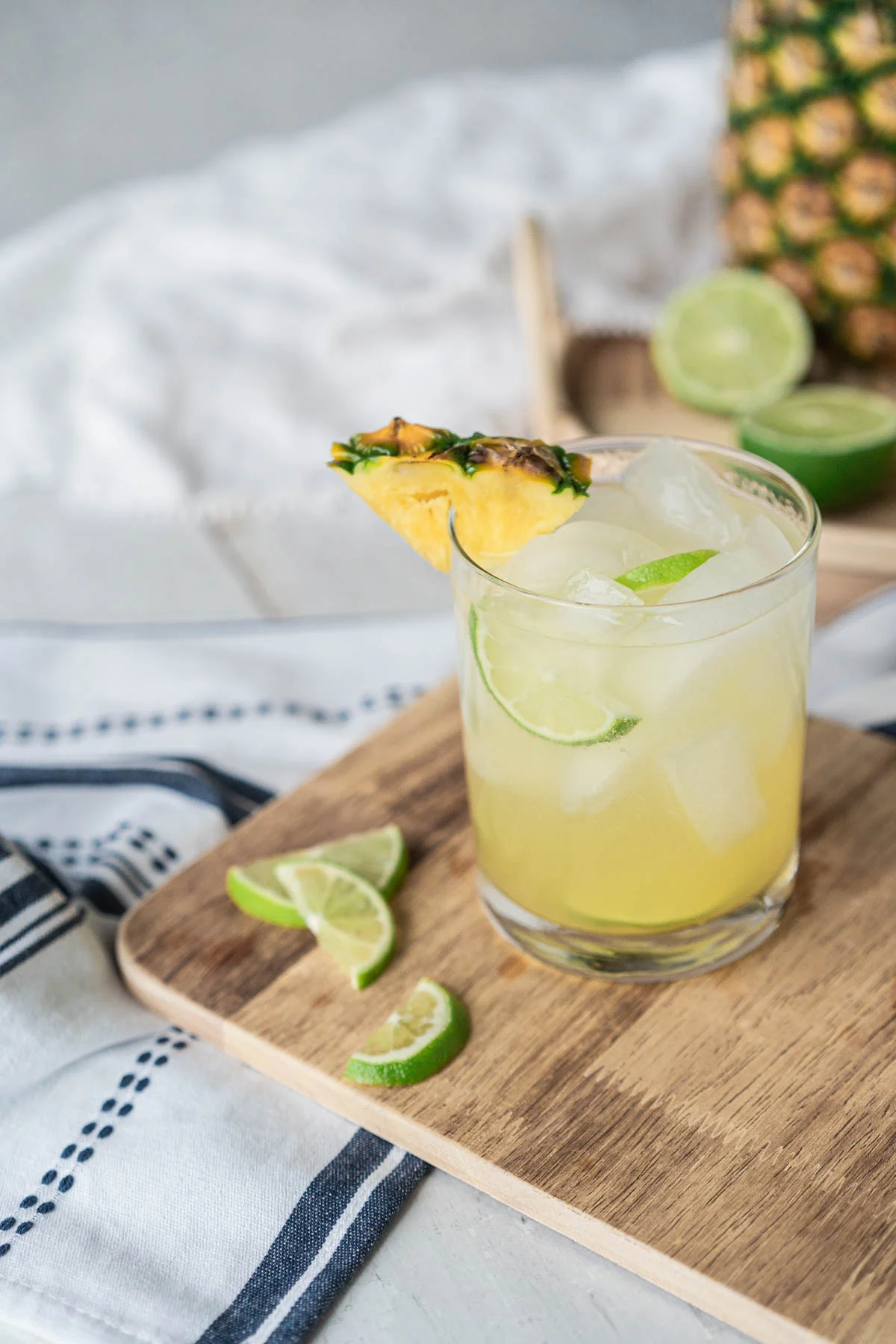 In the foreground, a rocks glass filled with ice and a light yellow drink, garnished with lime and pineapple wedges sits on a cutting board. Blurred in the background is a large pineapple and sliced limes