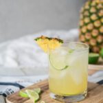 rocks glass filled with ice and a pale yellow drink garnished with lime and pineapple wedges