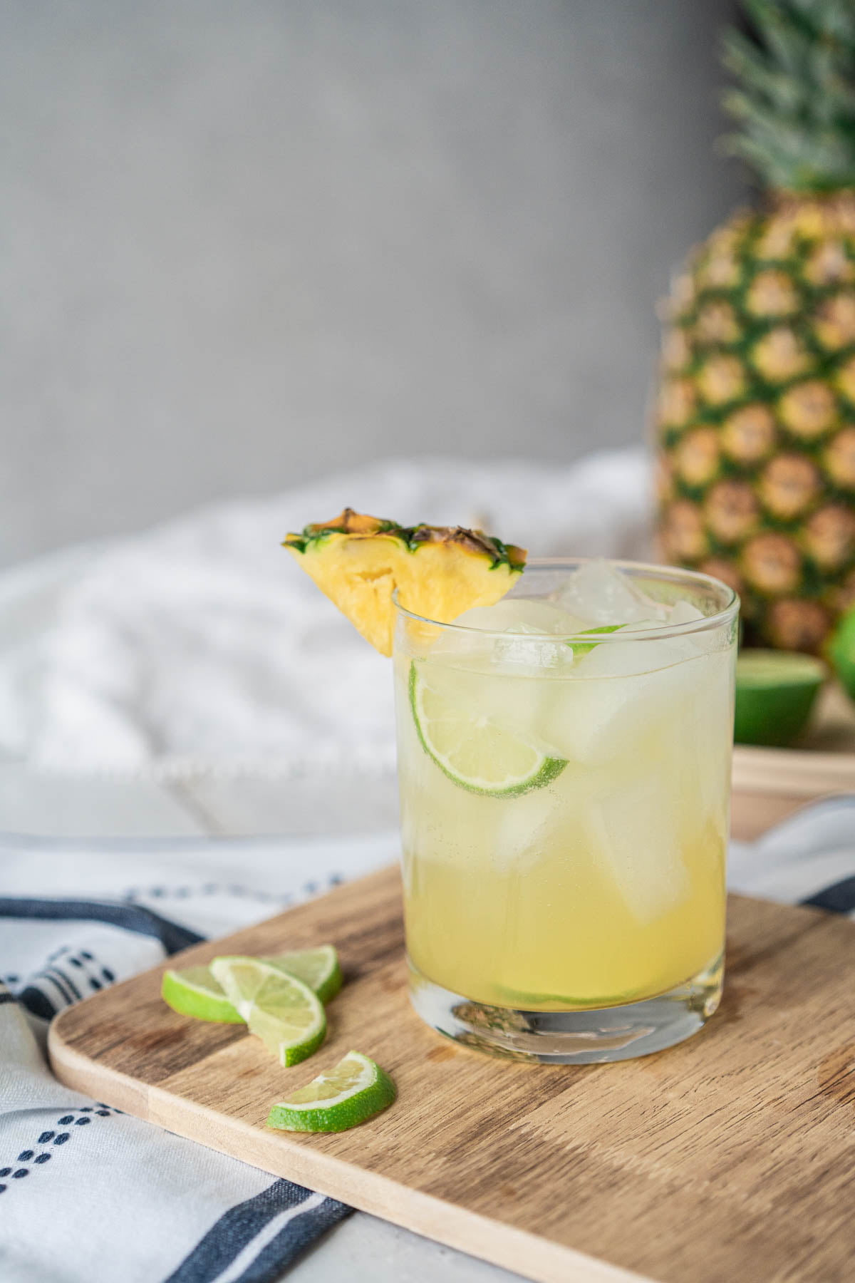 In the foreground, a rocks glass filled with ice and a light yellow drink, garnished with lime and pineapple wedges sits on a cutting board. Blurred in the background is a large pineapple and sliced limes