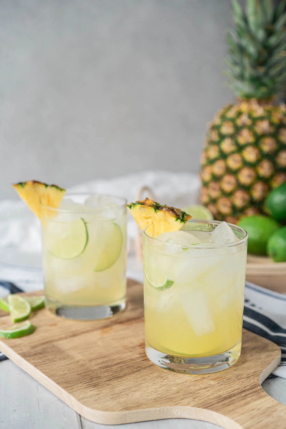 In the foreground, two rocks glasses filled with ice and a light yellow drink, garnished with lime and pineapple wedges sit on a cutting board. Blurred in the background is a large pineapple and sliced limes