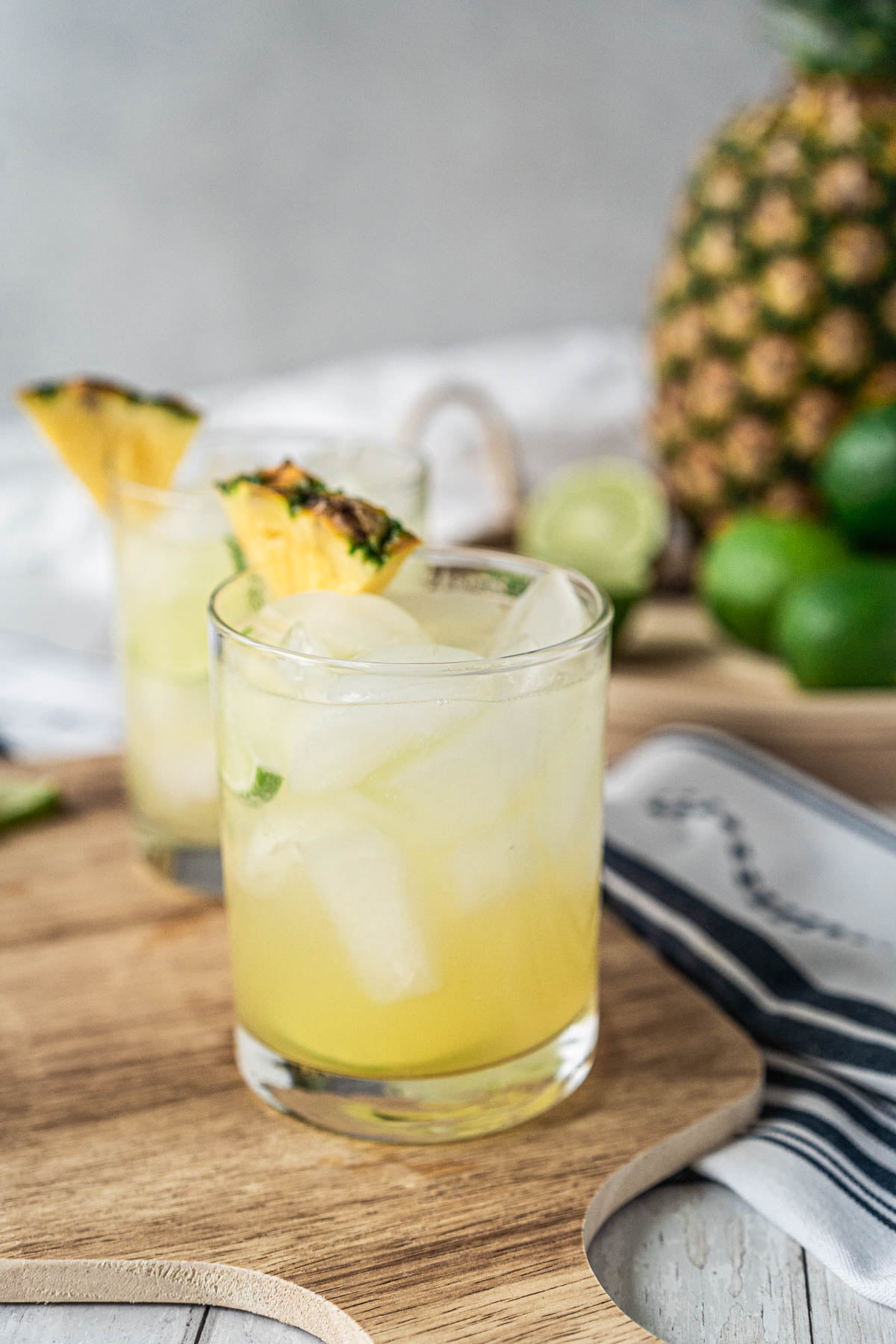 In the foreground, a rocks glass filled with ice and a light yellow drink, garnished with lime and pineapple wedges sits on a cutting board. Blurred in the background is another matching glass, a large pineapple, and sliced limes
