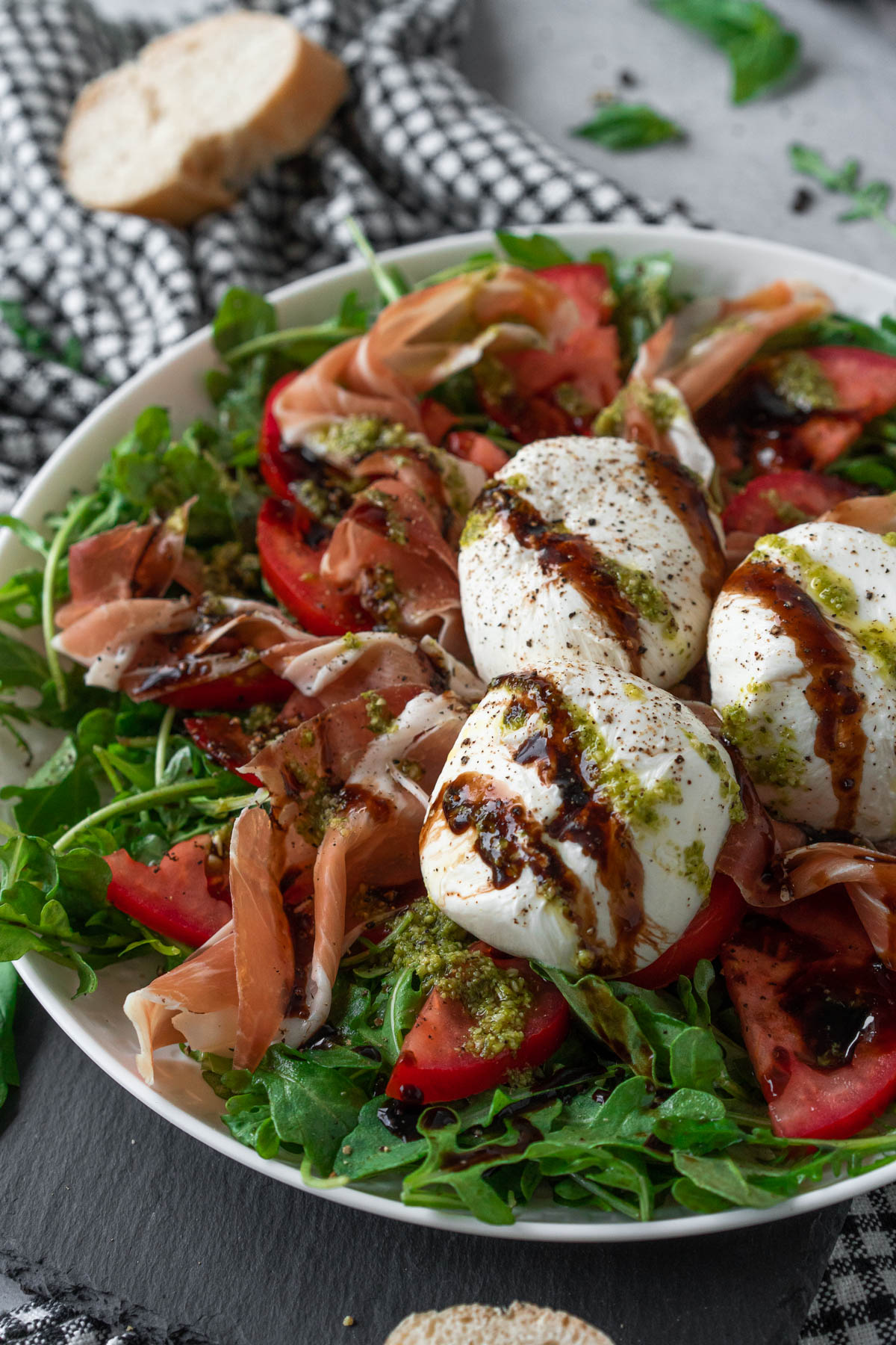 closeup shot of a white plate on a black and white checkered napkin; the plate is filled with an arugula salad topped with prosciutto ribbons, tomato slices, three burrata balls, and balsamic and pesto drizzle