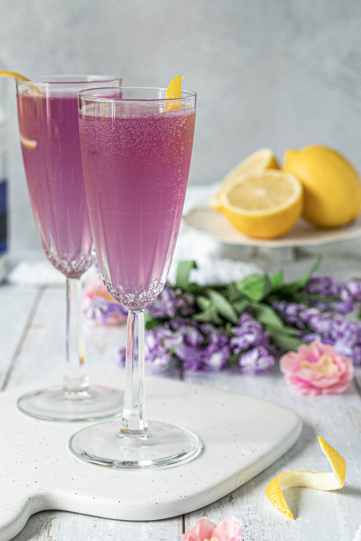two champagne flutes filled with a pale purple bubbly drink and garnished with lemon twists 