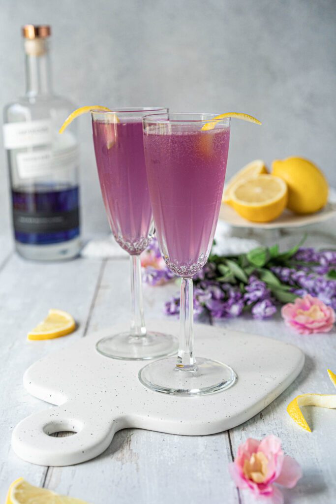 two champagne flutes filled with a pale purple bubbly drink and garnished with lemon twists