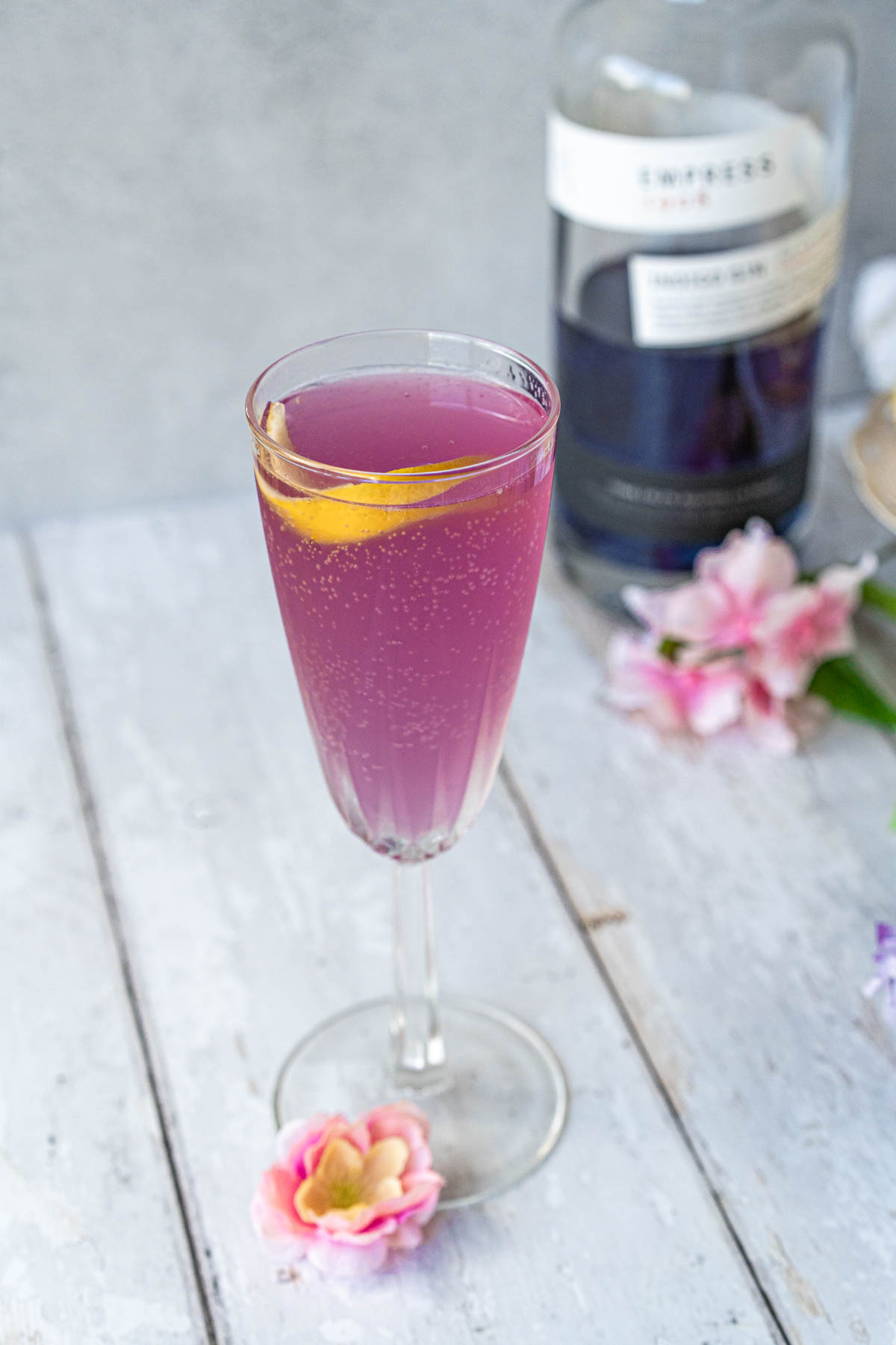 crystal champagne flute filled with a pale purple bubbling cocktail and garnished with a lemon twist; bottle of empress gin is blurred in the background