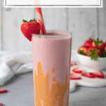 tall glass filled with a pink smoothie that is drizzled with peanut butter and garnished with a fresh strawberry; text overlay says 'high-protein strawberry PB smoothie'