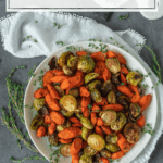 top-down view of roasted sliced carrots and Brussels sprouts garnished with fresh thyme on a white plate against a white napkin and gray tabletop with sprigs of fresh thyme in the background