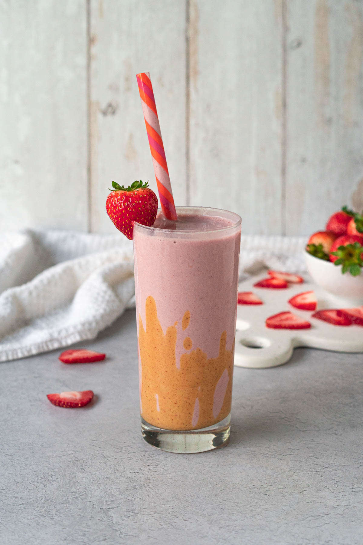 glass filled with strawberry smoothie garnished with peanut butter drizzle, fresh strawberry, and a pink and orange striped straw