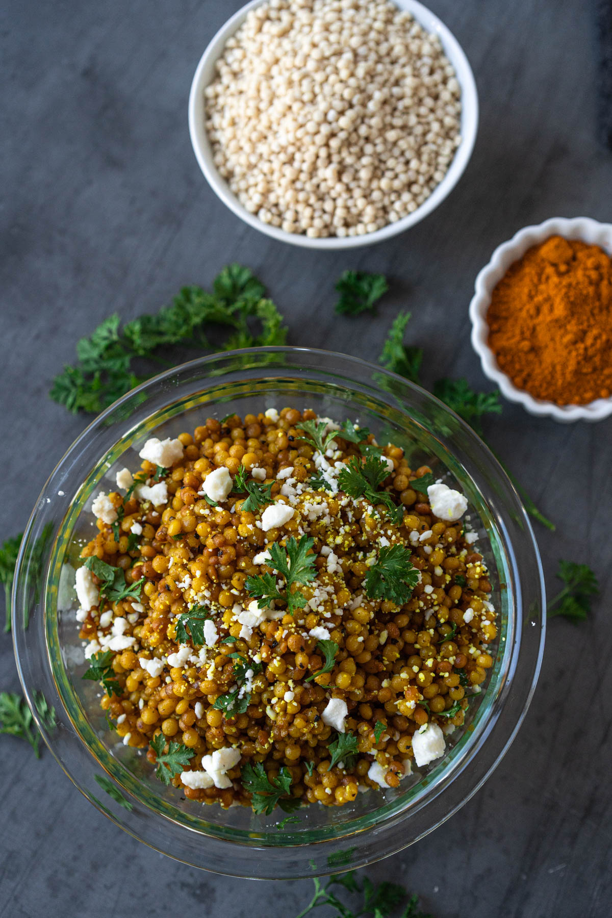Clear bowl filled with golden couscous, garnished with fresh parsley and crumbled feta. The bowl is on a gray surface that has fresh parsley scattered across it. Blurred in the background are two bowls – one with ground turmeric and the other with dry couscous