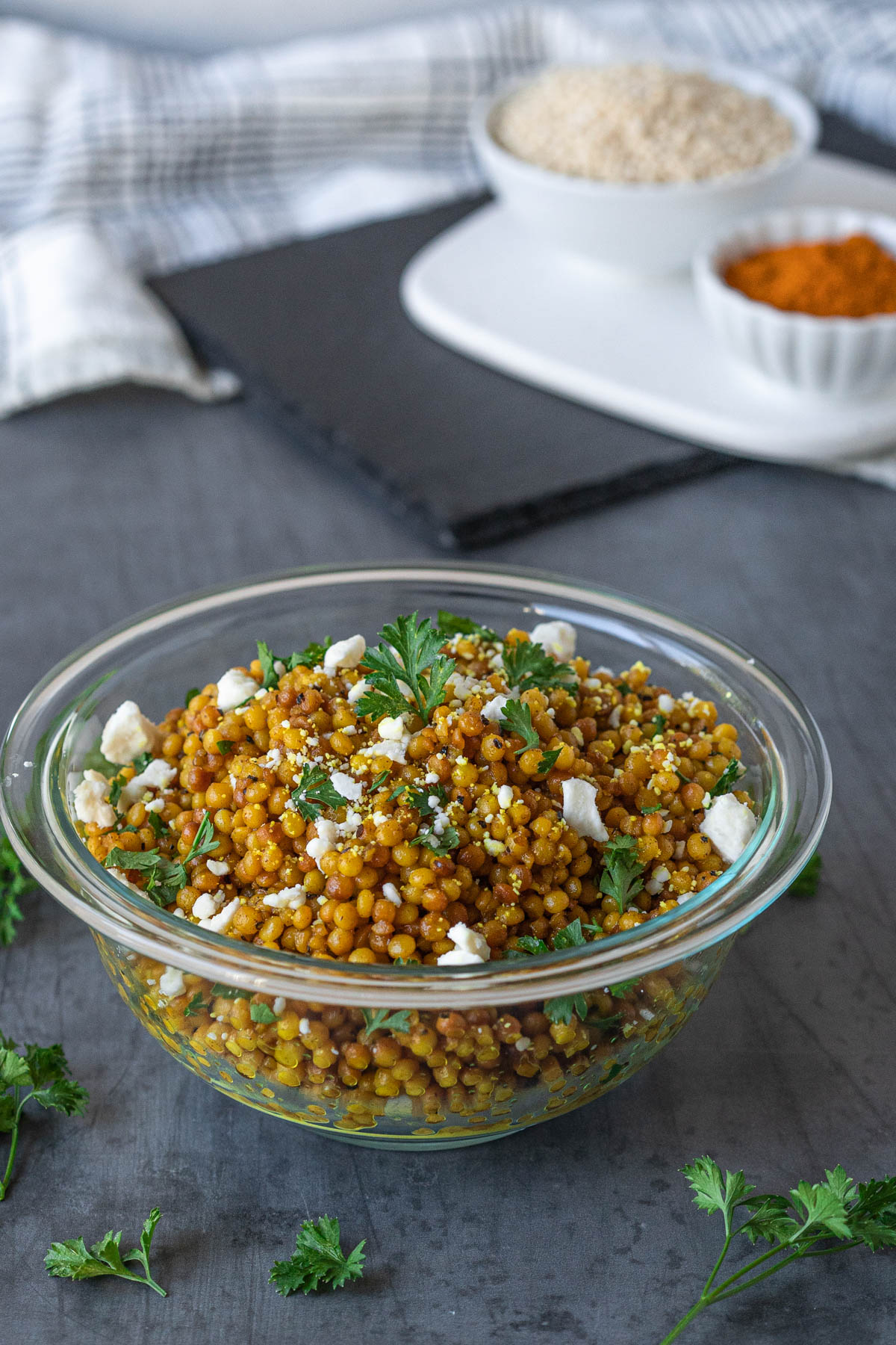 Clear bowl filled with golden couscous, garnished with fresh parsley and crumbled feta. The bowl is on a gray surface that has fresh parsley scattered across it. Blurred in the background, there are black and white serving platters with bowls of dry couscous and ground turmeric in them.