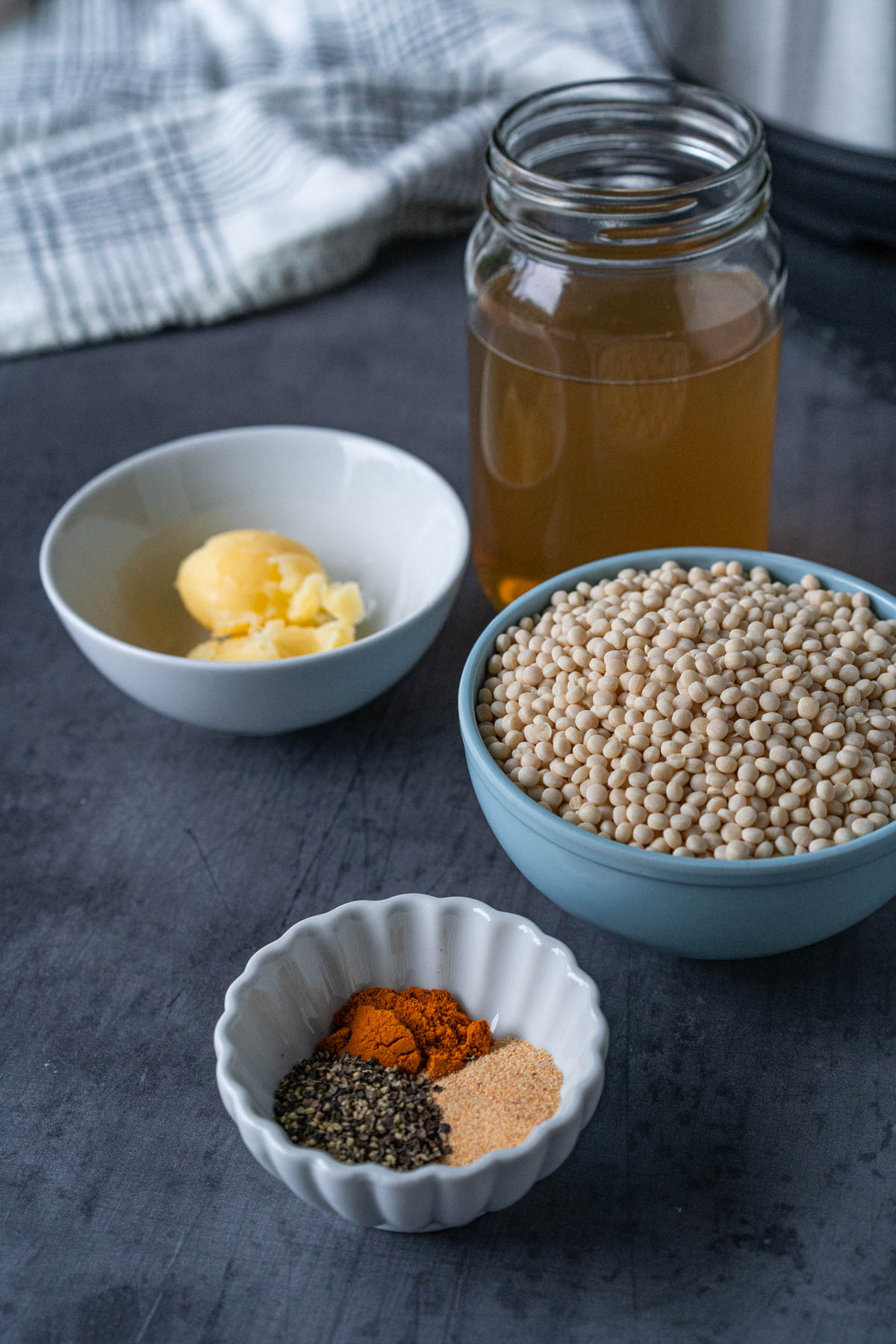 Side-view of spiced couscous ingredients – small bowl of garlic powder, ground turmeric, and black pepper, larger bowl with two tablespoons of ghee, a larger bowl with dry couscous, and a clear jar of chicken broth