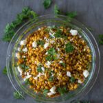 Clear bowl filled with golden couscous, garnished with fresh parsley and crumbled feta. The bowl is on a gray surface that has fresh parsley scattered across it.