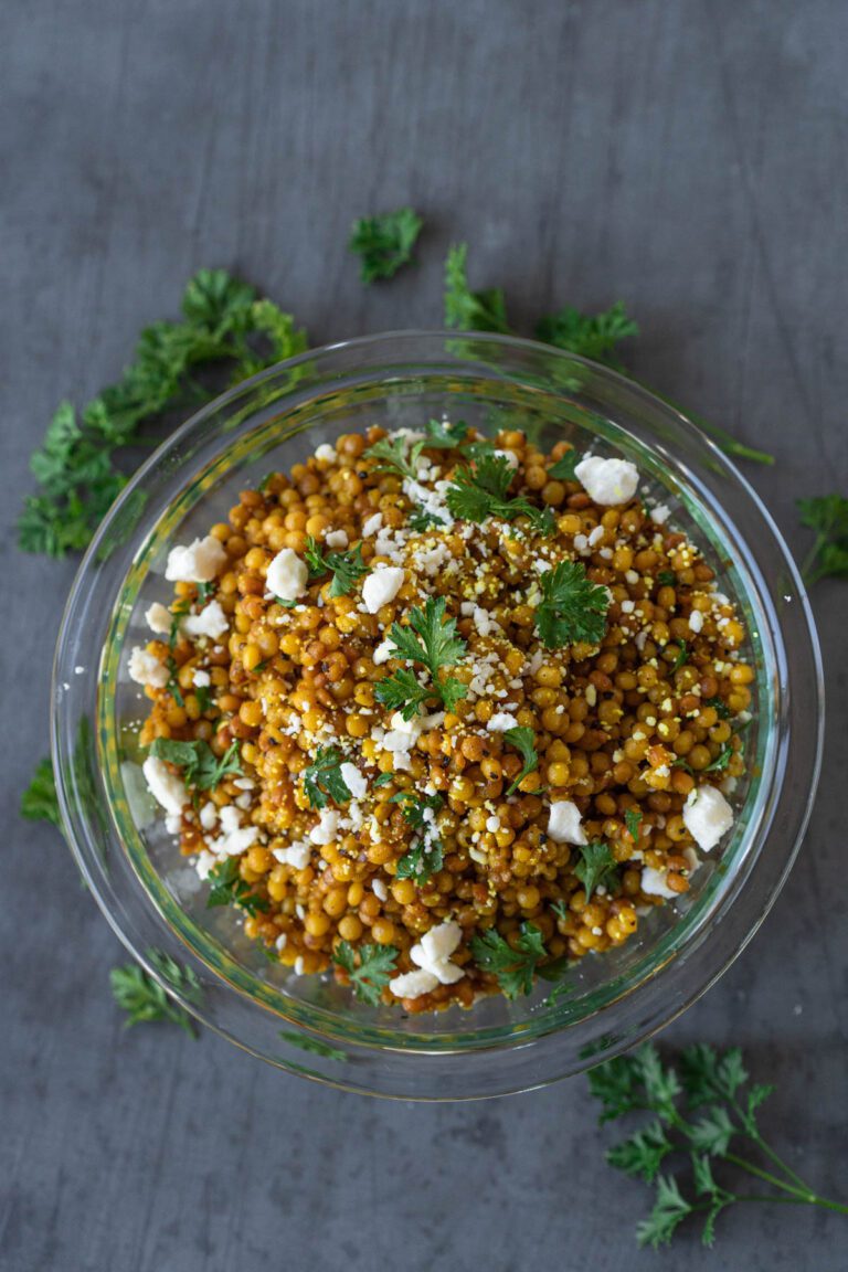 Clear bowl filled with golden couscous, garnished with fresh parsley and crumbled feta. The bowl is on a gray surface that has fresh parsley scattered across it.