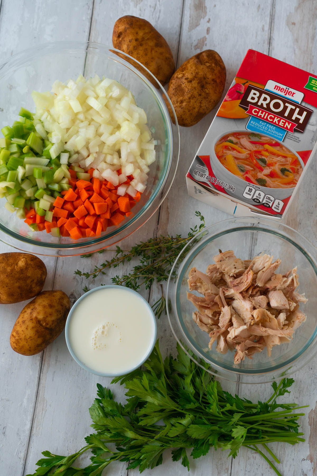 Top-down view of turkey and potato soup ingredients. From left to right - large, clear glass bowl of diced celery, onions, and carrots; four small russet potatoes; a cup of milk; fresh thyme, parsley, and rosemary; a small, clear glass bowl of shredded turkey; and a 48 oz carton of chicken broth