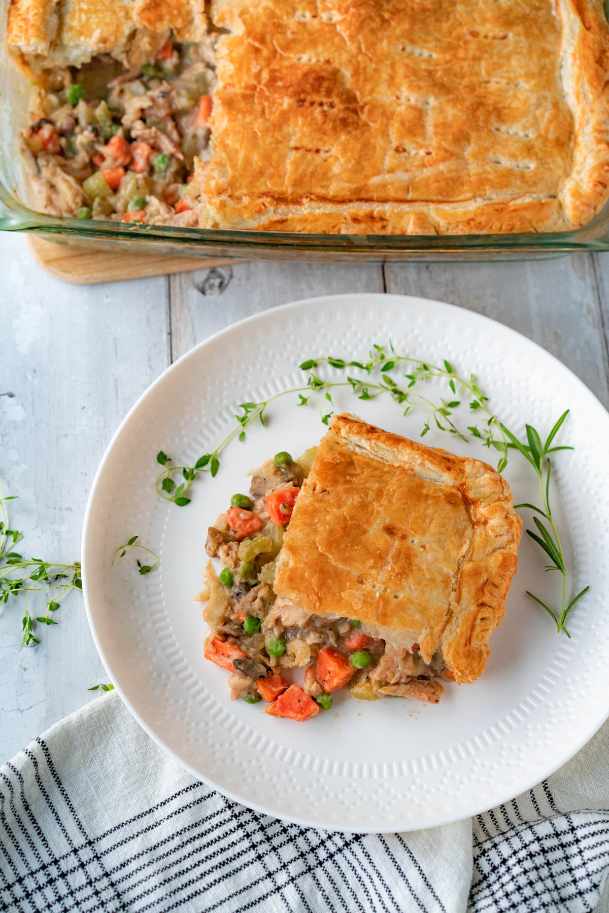 Top-down view of a rectangular slice of turkey pot pie on a small white plate, garnished with fresh thyme and rosemary. Above the plate is a baking dish of turkey pot pie with a slice missing. Below the plate is a white napkin with a black plaid pattern