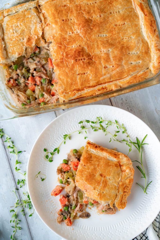 Top-down view of a rectangular slice of turkey pot pie on a small white plate, garnished with fresh thyme and rosemary. Above the plate is a baking dish of turkey pot pie with a slice missing.