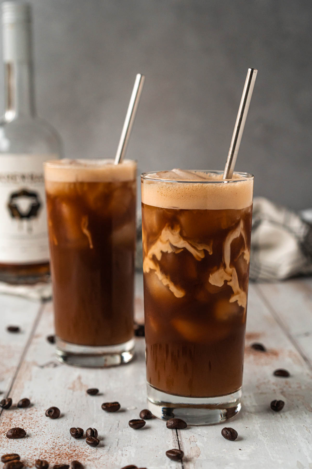 Side-view photo of two tall glasses filled with iced coffee and cream cascading down from the top. A bottle of peanut butter whiskey is blurred in the background