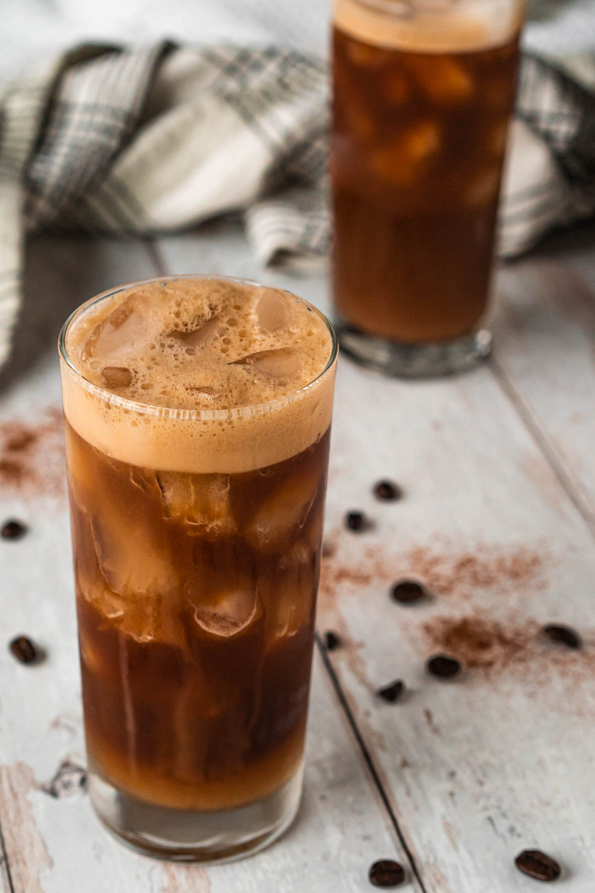 Closeup shot of a tall glass filled with iced coffee with foam on top and cream cascading down the side of the glass. Another glass of iced coffee is blurred in the background.