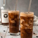 Side-view photo of two tall glasses filled with iced coffee and cream cascading down from the top. A bottle of peanut butter whiskey is blurred in the background