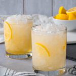two rocks glasses filled with crushed ice and a pale yellow drink with sliced lemon wedge garnishes