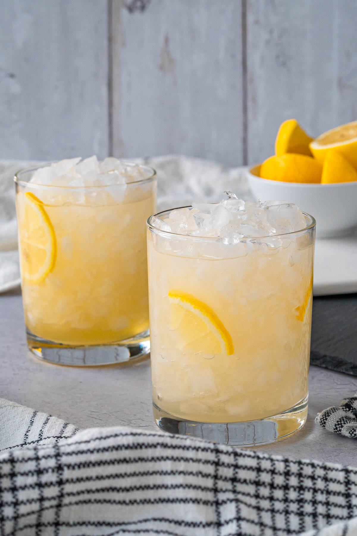 two rocks glasses filled with crushed ice and a pale yellow drink with sliced lemon wedge garnishes