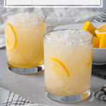 two rocks glasses filled with crushed ice and a pale yellow drink with sliced lemon wedge garnishes; a white text box overlays the top of the image and reads 'ginger beer mocktail with honey + lemon'