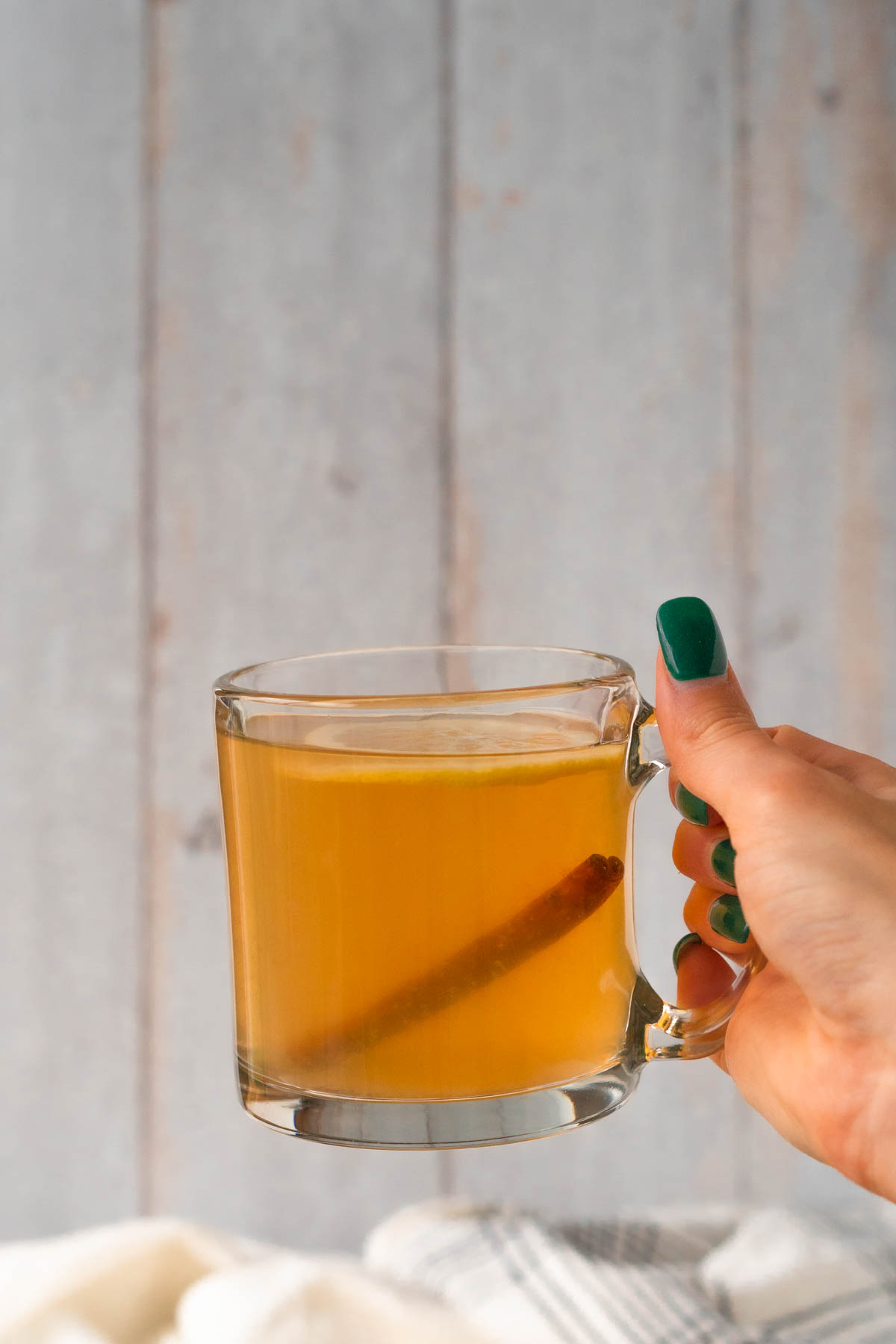 hand holding a clear mug filled with an amber liquid and topped with a lemon slice and cinnamon stick