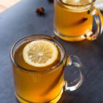two clear mugs filled with an amber liquid and topped with a lemon slice and cinnamon stick